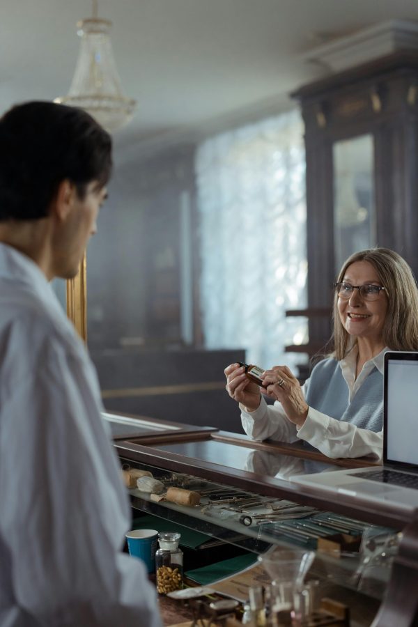 Senior woman pharmacist assisting a customer with a bottle in a vintage pharmacy.